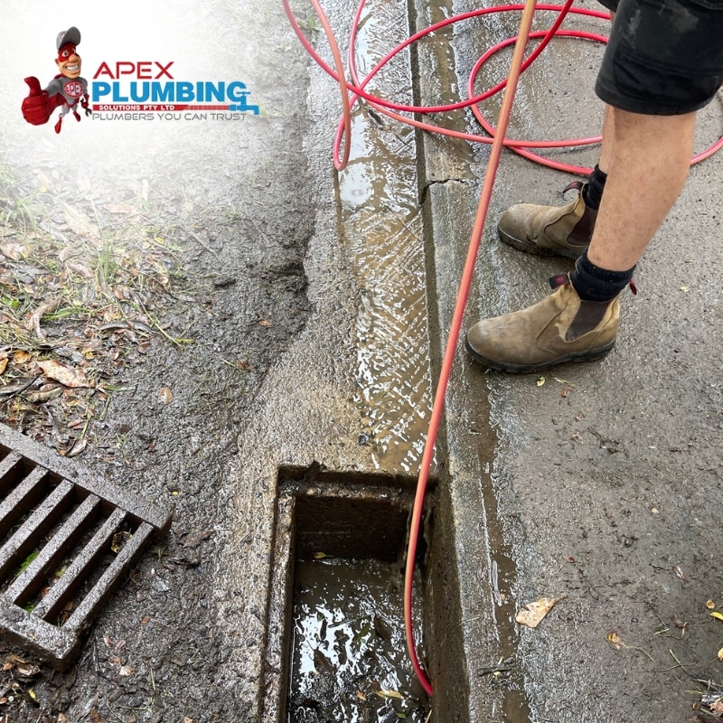 A man using a drain cleaner, which is a hose attached to a machine, to clear a clogged drain on a sidewalk.