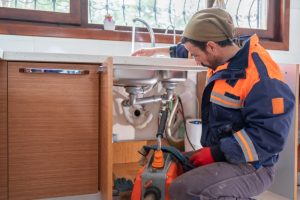 Professional plumber fixing a kitchen sink under a wooden countertop. The man is wearing a blue and orange work jacket and a beanie, using a plumbing snake machine. Kitchen sink dropped repair in progress with visible pipes and tools under the sink.