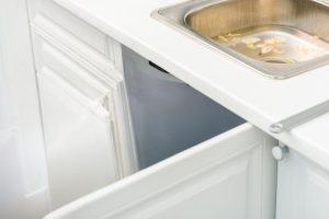 Kitchen sink dropped partially into the cabinet with white cabinets and countertops. The stainless steel sink is filled with water and vegetable peels, indicating a potential plumbing issue. Open cabinet doors beneath the sink reveal the extent of the problem.