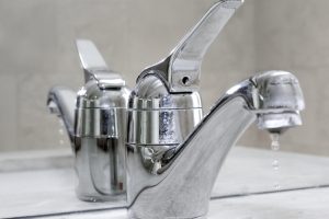 Close-up of a chrome bathroom faucet with tap dripping water, highlighting the issue of a leaky tap. The reflection of the faucet in the mirror emphasizes the water droplets, showcasing the need for repair.