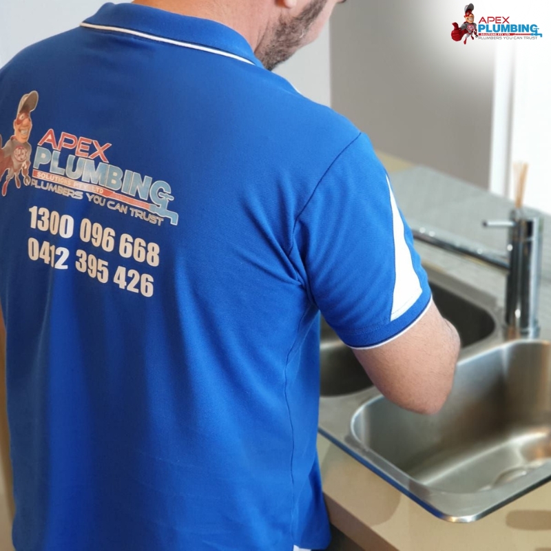 An Apex Plumbing professional providing plumbing services at a kitchen sink in the Eastern Suburbs of Sydney. The plumber, wearing a blue uniform with the company’s contact information displayed on the back, exemplifies reliable and trusted service in the region.