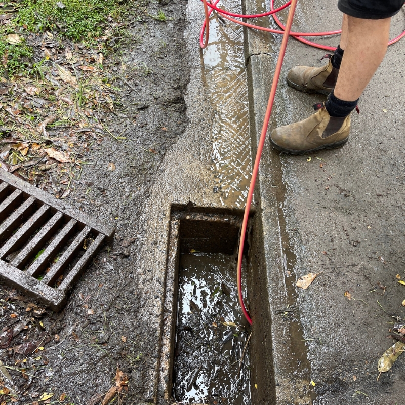 : A plumber for blocked drains is using a tool to clear a blocked drain on a muddy and wet roadside, with the drain cover removed and the area filled with debris and water.