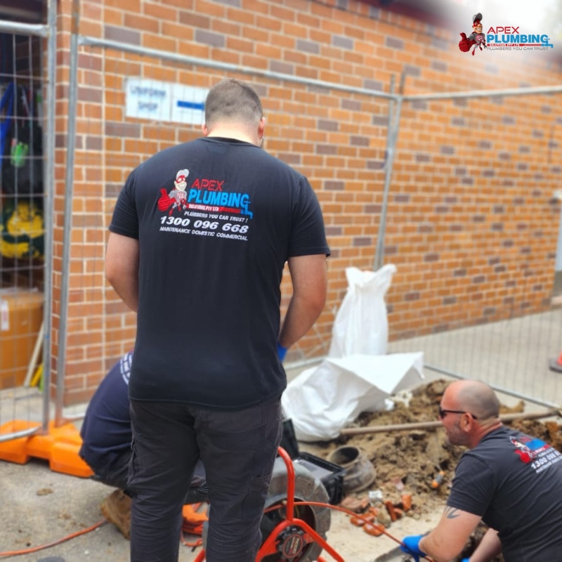 Plumber in Northern Beaches repairing a pipe outdoors, wearing branded Apex Plumbing shirts, showcasing professionalism and teamwork at a construction site with safety barriers.