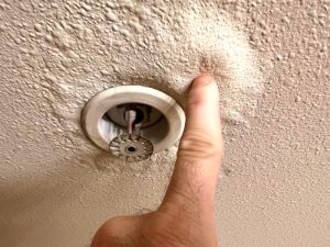 Person inspecting roof condensation leaks, pressing on a water-damaged ceiling near a fire sprinkler, showing visible swelling.