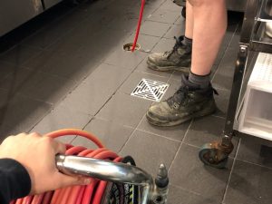 A close-up image of professionals doing jet blasting plumbing to clean floor drain in commercial kitchen.