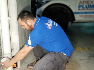 Plumber fixing a burst pipe under house next to white wall.