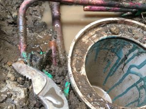 Close-up of muddy burst pipe under house with water and tools visible.