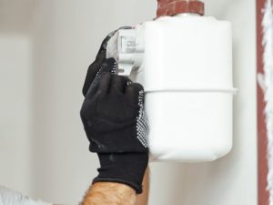 A technician wearing black gloves inspecting a potentially faulty gas meter installed on a white wall.