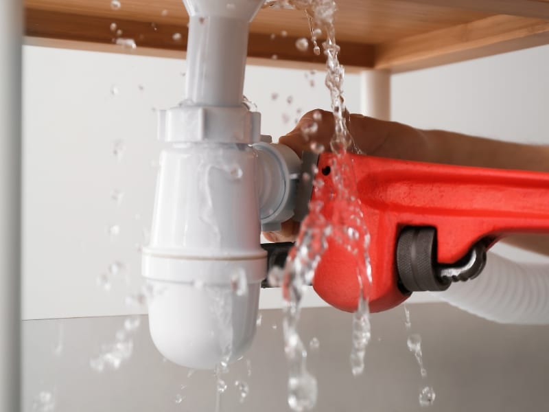 A leaky white pipe in an older home, being tightened with a red wrench, showing a common issue of burst pipes in older homes.