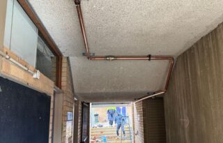 Copper gas pipe installed along a concrete ceiling in a building corridor during commercial gas fitting work, with tradespeople visible outside.