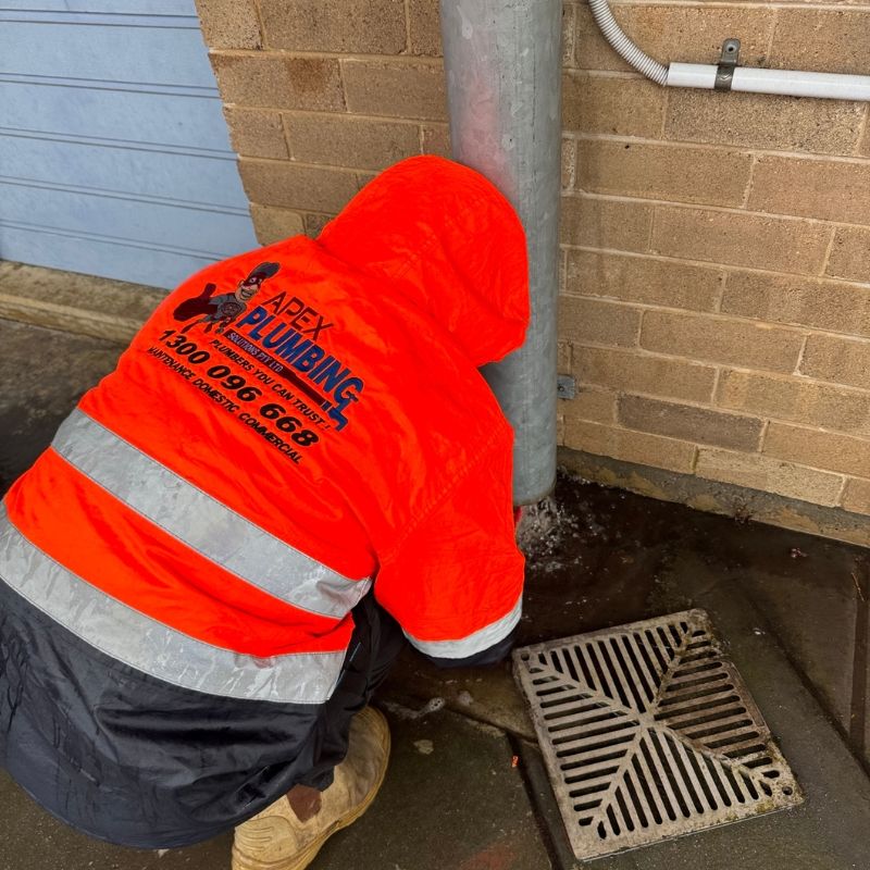 Apex plumber clearing an outdoor drain grate beside a brick wall