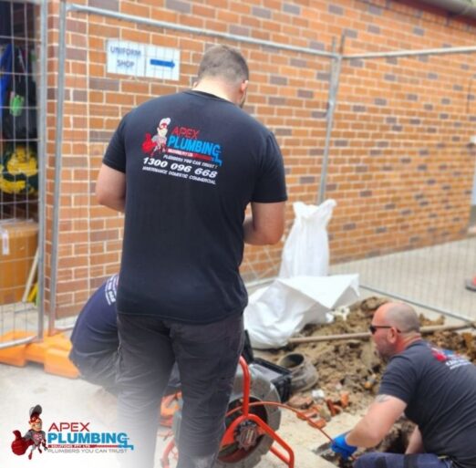 Licensed plumbers working beside an excavated trench to repair a blocked stormwater drain outside a brick building.