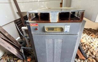 Outdoor gas hot water system unit with a rusted flue and vented metal casing installed beside a residential wall.
