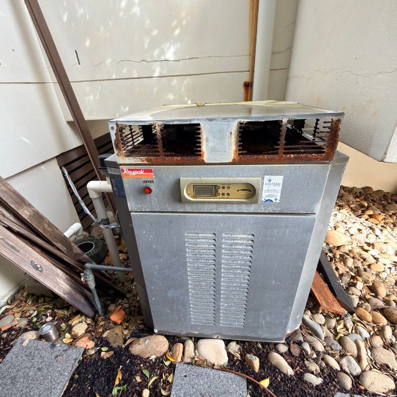 Outdoor gas hot water system unit with a rusted flue and vented metal casing installed beside a residential wall.