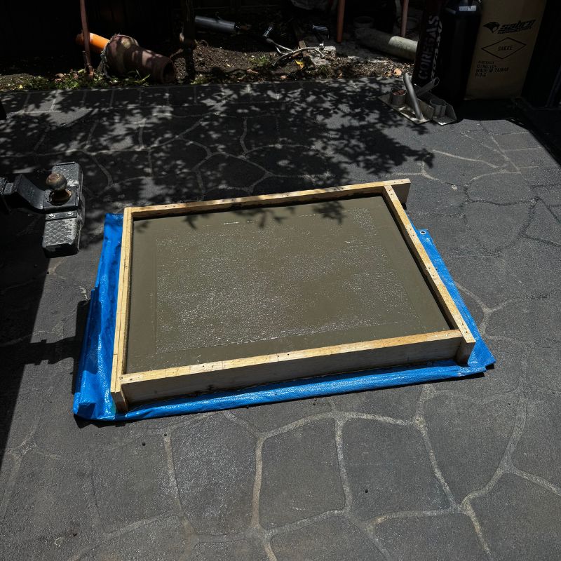 Temporary wooden formwork filled with wet concrete covering a blocked stormwater drain on a paved outdoor surface, preventing water flow.